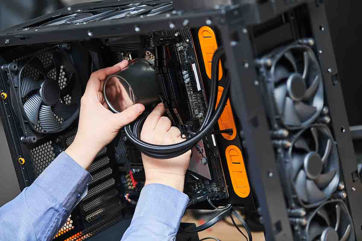 A person installs a circular cooling fan into a computer case. They are carefully handling the fan cables, with a focus on ensuring proper setup inside the chassis which also contains multiple fans and orange cable management accents. Visible parts include RAM slots and various connection ports on the motherboard.